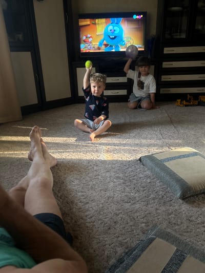 Two children are sitting on the floor in front of a television, with an adult present, possibly in a domestic setting. The TV is displaying a cartoon. The location is potentially in Klin, Russia, based on the presence of Russian children's programming, though this is not definitive without additional evidence.