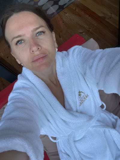 The image shows a close-up selfie of a woman wearing a white terrycloth bathrobe with a gold embroidered logo. The logo appears to be a horse with some text beneath it, which seems to read "ЦАРСКАЯ" and "АРХИВ". The woman has fair skin, blue eyes, and brown hair pulled back. She is wearing a delicate gold necklace and small pearl earrings. The background consists of wooden paneling, a red surface, and a light-colored pillow or cushion. The overall impression is of a relaxed, possibly spa-like setting. The image was taken indoors.