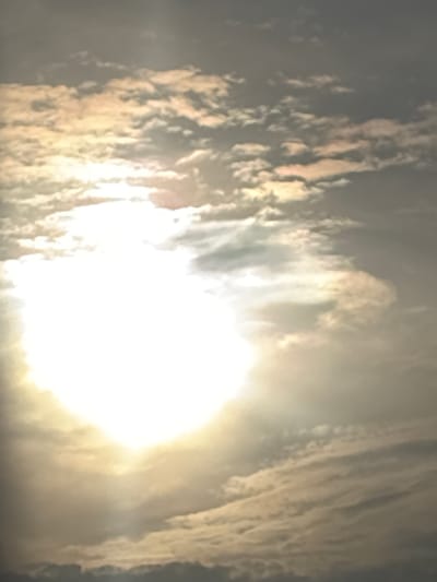 The image displays a dynamic sky scene, primarily composed of clouds and an intensely bright light source. The central and right portions of the frame are dominated by a powerful glare, indicating a direct view of the sun, which appears as a brilliant white, overexposed mass. Streaks of light emanate from this core, illuminating the surrounding cloud formations.

The clouds are varied in texture and shape, appearing as elongated, somewhat wispy, and textured bands across the sky. They are particularly noticeable on the left and right sides of the bright light. Many cloud sections are tinged with warm golden and soft yellow hues, suggesting the sun's light is either breaking through or reflecting off them. Other areas of the clouds are darker, providing depth and contrast to the brightly lit sections. The overall effect is a diffused, radiant atmosphere.

No people, man-made objects, or discernible text are visible in the image. The scene is a purely natural landscape focused on the sky. The intense brightness and golden tones suggest either a midday sun behind thin clouds or a morning/late afternoon sun at an angle. The provided context indicates the location as Kukorevo, Bulgaria, though no specific geographical features are present in the sky view itself.
