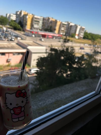 A person is holding a Hello Kitty themed glass filled with a frothy beverage. A black and white striped straw is inserted into the glass. The glass is positioned in the foreground, near a window. In the background, there are apartment buildings, trees, and parked cars, suggesting an urban environment. The scene appears to be an exterior view of a city, possibly Vidin, Bulgaria, given the architectural style of the buildings which are typical of Bulgarian socialist-era housing blocks. The sky is clear and blue, indicating a sunny day.