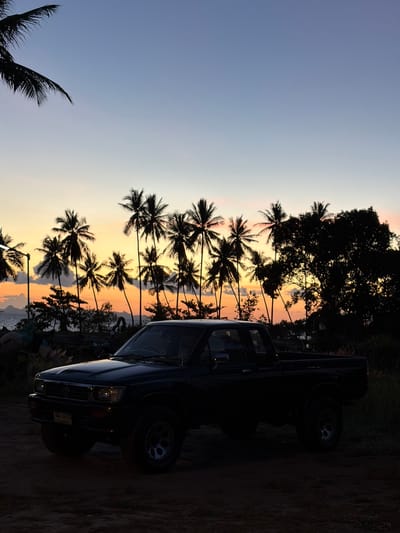 In Baan Taling Ngam, Thailand, a dark-colored pickup truck is parked on a dirt road at dusk. The truck is positioned in the foreground, facing left, with its headlights and grille visible. Behind the truck, a line of silhouetted palm trees stretches across the horizon, interspersed with other trees and foliage. The sky above displays a gradient of colors, transitioning from a pale blue at the top to warm orange and pink hues near the horizon, indicating sunset. In the distance, the ocean is visible, with faint outlines of islands or landmasses on the horizon. The overall scene evokes a tropical, serene atmosphere.