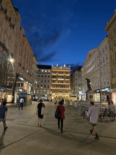 This image captures a street scene in Vienna, Austria, at dusk. The architecture is characteristic of the city, with grand, ornate buildings lining a wide pedestrian plaza. The sky is a deep, dark blue with scattered clouds, indicating twilight. The buildings are illuminated, highlighting their classical facades, balconies, and windows. Storefronts are visible along the street level, with some illuminated signs, including one for Louis Vuitton. Several people are walking in different directions across the plaza. In the foreground, on the right, a man in a light blue shirt and khaki shorts is walking away from the camera. To his left, a woman in a dark dress and a light-colored tote bag is also walking away. Further left, a person in a dark blue shirt and grey pants is walking towards the left. In the middle ground, there are more people walking, and a statue on a pedestal stands prominently in front of a large white building. Bicycles are parked near the statue. The ground is paved with large, light-colored stone slabs. The overall impression is of a lively urban environment transitioning from day to night, with historical architecture providing a rich backdrop.