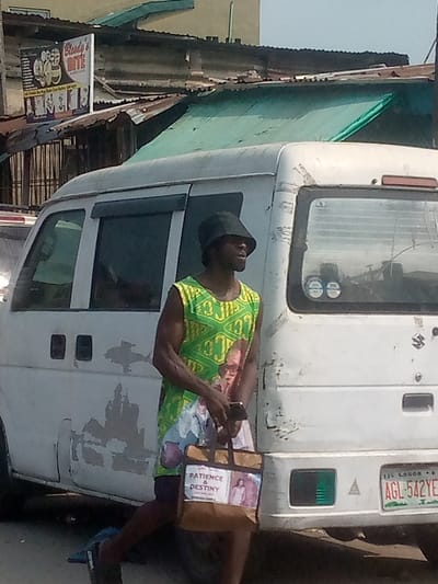 A man with a dark complexion wears a black hat and a green tank top with an image on the front. He carries a shopping bag with the words "PATIENCE & DESTINY" printed on it, along with pictures. In his other hand, he holds a smartphone. The man stands next to a white van with peeling paint. The vehicle's license plate, partially visible, indicates registration in Lagos. Buildings with corrugated iron roofs form the background. A sign for "Blusty's Bite" is visible in the upper left corner.