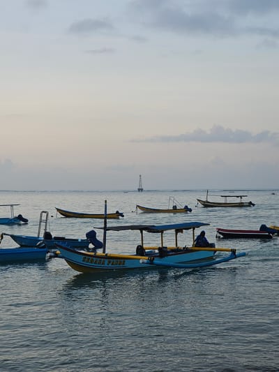 This is a photograph of a serene ocean scene, likely taken at a beach in Denpasar, Indonesia, possibly during sunrise or sunset given the soft, diffused light. The image is dominated by a calm, rippling sea that reflects the muted colors of the sky, which transitions from a pale blue at the top to a warmer, almost peachy hue towards the horizon. A few wispy clouds are scattered across the sky.

In the foreground and middle ground, numerous traditional Balinese outrigger boats are anchored or drifting gently on the water. These boats are of various sizes and colors, predominantly white, yellow, and blue, with some featuring protective canopies. One prominent boat in the center of the frame is light blue with yellow trim and has the text "SEDANA PANDE" painted on its side. A lone figure, wearing a blue hooded garment, is seated inside this boat, seemingly observing the surroundings. Other boats are scattered throughout the scene, with some closer to the viewer and others further out.

In the far distance, on the horizon, a tall, skeletal structure, possibly an oil rig or a navigation tower, stands out against the sky. The overall atmosphere is tranquil and picturesque, suggesting a peaceful moment by the coast.