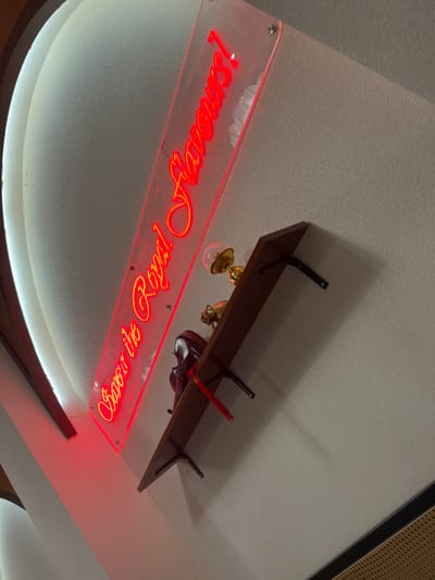 An upward-angled, tilted photograph captures an interior wall in the Chongqing High-tech Zone, China, featuring a red neon sign and a mounted shelf. The central element is a red neon sign, illuminated brightly, reading "Savour the Royal Flavours!" in a flowing script, mounted on a clear acrylic panel. The sign is affixed to a light-colored, textured wall. Below the sign, a dark wooden shelf is horizontally mounted to the wall with two visible black metal brackets. Objects arranged on the shelf include a golden, ornate lamp with a spherical top, and a dark, possibly red-brown, decorative box or container adorned with a red tassel. To the far left, a curved architectural element, possibly a ceiling cove or recessed archway, is illuminated from within, creating a bright white glow along its edge and bordered by dark wood trim. The wall exhibits a consistent stippled or speckled texture. A shadow from the shelf is cast on the wall below it. In the bottom right corner, a section of a woven, possibly rattan or wicker, panel with a dark frame is partially visible. No people are present.