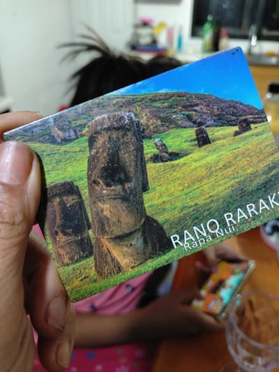 The image shows a close-up of a postcard featuring the Rano Raraku quarry on Easter Island (Rapa Nui), Chile. The postcard depicts several Moai statues in a grassy landscape under a blue sky. A person's hand is holding the postcard, obscuring part of the scene. In the background, out of focus, a child is seen holding a smartphone. A glass with some food is also visible in the foreground. While the postcard itself represents a location in Chile, the immediate scene is not in the city of Arica.