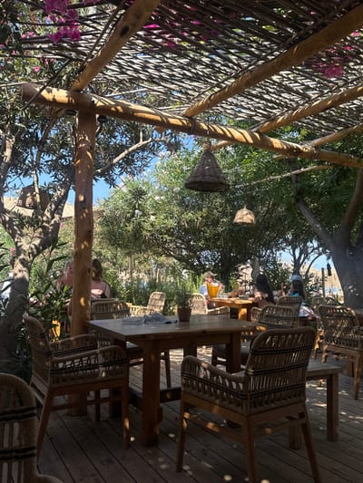 The image depicts an outdoor dining area in Lambi, Greece, under a wooden pergola providing shade. The pergola's roof is constructed from interwoven branches or reeds, with visible pink bougainvillea flowers growing over it. Two cone-shaped, woven pendant light fixtures hang from the pergola's beams. The ground is a wooden deck.

Multiple wooden tables and woven rattan or wicker chairs are arranged throughout the space. In the foreground, a square wooden table is visible with an ashtray and a folded dark cloth on its surface, surrounded by four empty woven chairs.

In the mid-ground and background, several individuals are seated at other tables. To the right, one person wearing a cap is seated at a wooden table with a yellow bucket, possibly containing ice. To their left, two individuals with dark hair are seated at another table. Further to the left, partially obscured by foliage and a wooden pillar, two additional people are partially visible, one with their back to the viewer wearing a dark top, and another's face partially seen.

The area is surrounded by dense green trees and shrubs, providing a natural, shaded environment. Sunlight filters through the pergola and foliage, casting dappled shadows.