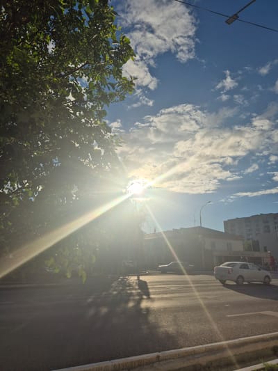 The image depicts a bright, sunny day in Kokshetau, Kazakhstan. The scene is a street with a sidewalk and a road. The sun is shining brightly, creating lens flare and highlighting the clouds in the blue sky. In the foreground, there is a leafy green tree. Beyond the tree, there are buildings, including a multi-story building and a smaller one with a sign. Several cars are parked on the side of the road, and one is driving away. The road appears to be asphalt with a yellow line marking the edge. The time of day is clearly daytime, and the weather is sunny and clear with scattered clouds. The overall mood is bright and cheerful due to the strong sunlight. There is some visible text on a sign on the smaller building, but it is not legible.