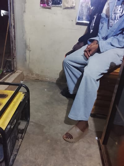 In Durumi, Nigeria, a close-up shot shows the lower bodies of two individuals sitting. The person in the foreground, wearing a light blue denim jacket and light gray sweatpants, is seated on a wooden structure. They have their hands clasped in their lap and are wearing a light brown slide sandal on their right foot. Their left foot is partially visible, wearing a dark shoe. To the left, a yellow generator with black trim is positioned on a concrete floor. In the background, a plain wall with some water damage and debris is visible. Above the wall, several items are hanging, including a framed picture or mirror. The scene appears to be indoors or in a sheltered outdoor area with dim lighting.