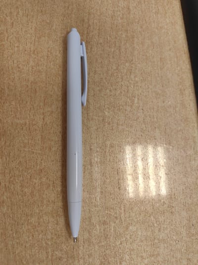 The image shows a white pen lying on a light brown, wood-like surface. The pen is positioned vertically in the left half of the frame, with its clip facing upwards and towards the left. The pen appears to be a standard ballpoint pen. The surface it rests on has a textured, grainy appearance, typical of laminate or wood. To the right of the pen, a shadow is cast, with distinct vertical lines of light shining through, suggesting the presence of blinds or a similar light-filtering object outside the frame. The lighting conditions indicate it is daytime. The overall scene is simple and focuses on the pen as the main subject. There is no visible text in the image. The location context of Ulyanovsk, Russia, does not provide specific visual cues within the image itself.