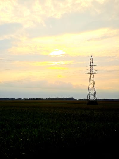 An electricity pylon stands in a rural field at sunset in Pawłów, Poland. The sky is a hazy yellow-orange with scattered clouds, and the sun is partially obscured by them. The pylon is a tall, lattice-steel structure with wires stretching across the frame. In the foreground, a dark field of crops, possibly corn or wheat, occupies the lower half of the image. A dark, tree-lined horizon is visible in the middle ground. The overall impression is one of a serene, agricultural landscape at dusk.