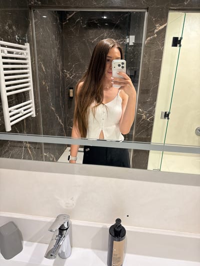 A woman is taking a selfie in a modern bathroom. She is in the center of the frame, holding a phone in her right hand, capturing her reflection in a large mirror. She has long, dark brown hair that falls over her shoulders and is wearing a white, ribbed, sleeveless top with a V-neck. The top has a few buttons at the neckline. On her left wrist, she wears a white smartwatch.
The bathroom features dark marble walls with white veining, creating a luxurious and contemporary feel. A white towel rack is visible on the left side of the frame. To the right of the woman, a white, textured wall dominates the space. A chrome faucet with a single handle is mounted on this wall, with water droplets visible on its surface. Below the faucet, a small gray soap dish sits on the counter. A large black pump bottle of what appears to be shampoo or body wash is mounted on the wall to the right of the faucet.
The lighting suggests it is daytime, possibly indoors. The overall mood is casual and personal, typical of a mirror selfie. The scene is set in a bathroom, likely in a hotel or a modern residential space. No discernible text is visible in the image, except for what might be on the label of the black pump bottle, which is too blurry to read. The location is Hisarya, Bulgaria.
