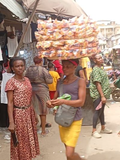 A crowded outdoor market scene depicts several individuals amidst various stalls and goods. A woman in a leopard-print dress and black belt stands in the foreground. Another woman walks with a stack of packaged items balanced on her head, wearing a gray tank top and yellow skirt, with a gray messenger bag. A third woman in a green patterned shirt and black leggings stands to the right. Other shoppers and market-goers are seen in the background, adding to the bustling atmosphere of the market.