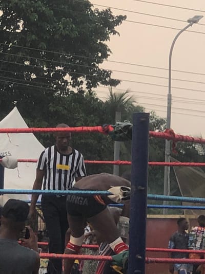 The scene depicts an outdoor professional wrestling event in Alor, Nigeria. In the foreground, a dark-skinned male wrestler, wearing black trunks with "JUNGA" printed in white on the rear, and red and white leg wraps, is bent over at the waist with his back to the viewer, positioned inside a wrestling ring defined by red and blue ropes and a prominent blue corner post. Behind him, a dark-skinned male referee in a black and white vertical striped shirt featuring a central horizontal yellow stripe and dark trousers stands facing into the ring. To the immediate left, a spectator in a dark shirt and black cap is visible from the shoulders up, holding a mobile phone in a recording or photographing stance. The background features lush green foliage from numerous trees, a partial view of a white tent on the left, multiple overhead power lines, and a tall street light with a curved pole on the right, all under an overcast or hazy sky.