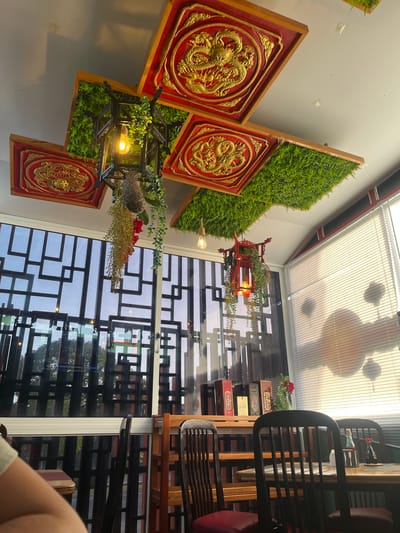 This image captures an interior view of a restaurant with an Asian-inspired theme. The foreground features decorative wall hangings with intricate golden dragon reliefs set against a red background, framed in wood. These are interspersed with artificial green foliage, creating a vertical garden effect. A stylized lantern with greenery and a lightbulb hangs in the center.
The background reveals a large window with a geometric, dark grid pattern overlaying the glass, offering a view of the outside. Through the window, trees and more of the restaurant's interior with tables and chairs can be seen. The ceiling is a translucent material with parallel lines, possibly a skylight or a covered section, with circular shadows cast from above, suggesting ambient lighting or decorative elements.
The image is taken from a low angle and rotated, giving a dynamic perspective. The lighting appears to be artificial, with warm tones from the lightbulbs, and natural light filtering through the ceiling. The overall impression is of a contemporary restaurant interior, likely open during the day or early evening, with a focus on decorative elements and ambiance. No people are clearly visible, and no text is discernible. The setting suggests a dining establishment, possibly in Porlamar, Venezuela, given the provided location context, with a design that aims to evoke a sense of tradition and nature.