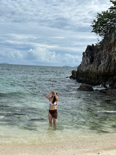 A person stands in the shallow, clear water at the edge of a sandy beach. The person is wearing a bikini, with a pink and white polka dot top and a brown bottom. They are facing towards the water and have their hands up near their head, obscuring their face. The water is calm with gentle waves lapping the shore. To the left of the person, a large, rocky cliff juts out, with some greenery growing at its top. The sky above is cloudy, with patches of blue visible. The scene appears to be set in a tropical location, possibly during the day. The mood seems relaxed and tranquil, with the person enjoying the water. There is no visible text in the image.