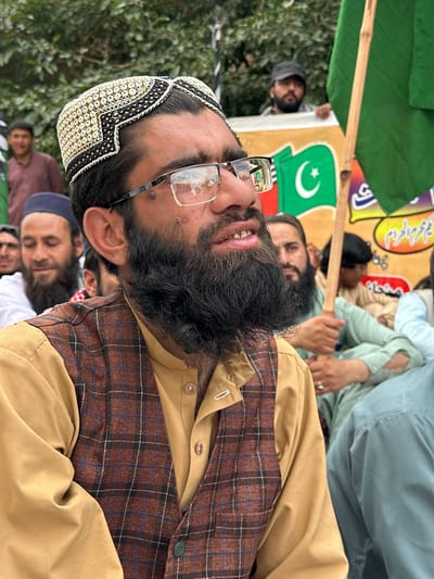 The image shows a close-up of a man wearing glasses, a traditional cap, and a plaid vest over a tan shirt. He has a beard and is looking to the right. In the background, other people are visible, some holding flags and banners, suggesting a public gathering or rally. One banner displays a green flag with a white crescent and star, likely the flag of Pakistan. Another person in the background is holding a green flag. The overall impression is of a political or religious event in an outdoor setting. The presence of the Pakistani flag suggests the location is within Pakistan. Given the context of similar events, and that Quetta City Tehsil is a significant administrative and urban area in Balochistan, Pakistan, this scene could plausibly be taking place in Quetta City Tehsil.