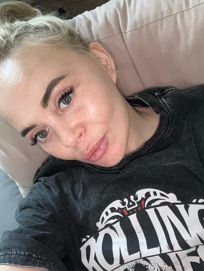 A woman is lying on a pillow, facing the camera. She has fair skin, blonde hair pulled back, and green eyes. She is wearing dark-colored shirt with a print on it. The background consists of a light-colored pillow and a grey surface. It is not possible to determine the location as Высокий, Russia from the image.