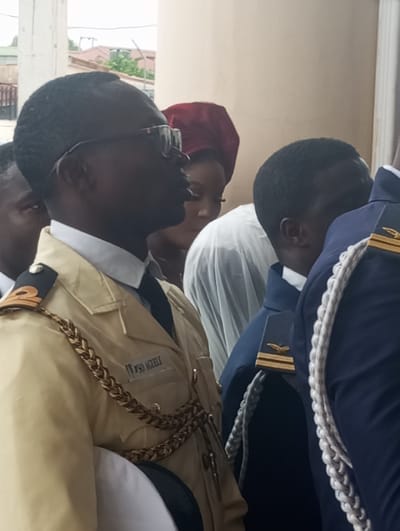 The image shows a close-up of several people in formal attire, possibly at an event in Barden Yana, Nigeria. In the foreground, a person wearing a light-colored uniform with gold braiding and epaulets with gold insignia is visible from the chest up. The uniform has a dark tie and a white collar. The name "VSO AGELLF" is visible on the uniform. To the right of this individual, another person is seen wearing a dark blue uniform with white braided cords and gold insignia on the epaulets. Further to the left and in the background, other individuals are partially visible. One person has a dark head covering, and another is seen wearing glasses. The background appears to be an interior or covered area with a pale wall and architectural elements suggesting a building, possibly a church or event hall, with hints of outdoor greenery and buildings visible through an opening. The lighting is diffused, indicating an overcast or indoor setting.