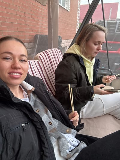 Two women are reclining on a cushioned swing or hammock outdoors. The woman in the foreground is looking at the camera and smiling. She is wearing a black jacket over a gray plaid shirt and a white t-shirt. A partial view of the second woman is visible. She is wearing a black jacket over a yellow hoodie and white pants and is using chopsticks. The background includes a brick building, foliage, and a partial view of a metallic frame. It is daytime. The location is possibly Ogryzkovo, Russia.
