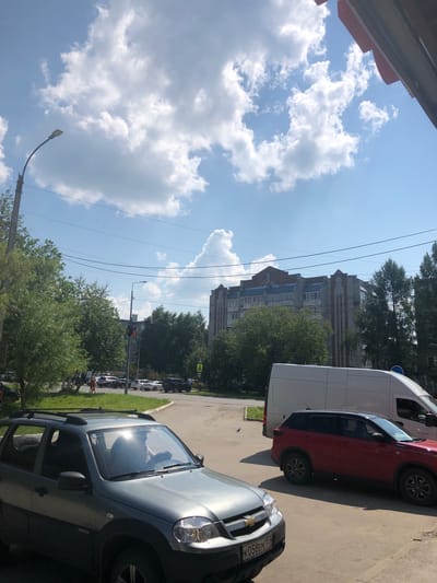 The image depicts a street scene in Solikamsk, Russia, on a sunny day with scattered clouds. In the foreground, a gray SUV is parked, with a driver visible behind the steering wheel. To the right, a red SUV is parked, and a white van is partially visible. In the background, there is a multi-story apartment building with a gray brick facade and red decorative elements. Trees line the street and sidewalks, and power lines are strung across the sky. Several other vehicles and pedestrians are present in the middle ground, suggesting a moderately busy urban environment. The road surface appears to be asphalt.