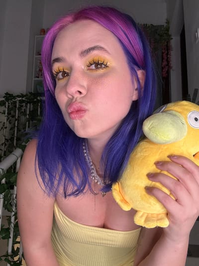 A person with purple and pink hair is holding a yellow Psyduck plush toy. They are wearing a yellow top and a pearl necklace. Their makeup includes yellow eyeshadow and glitter around the eyes. The background includes greenery and a shelf with items on it. The image appears to be taken indoors. The location is not discernible as Zagreb, Croatia based on the given image.