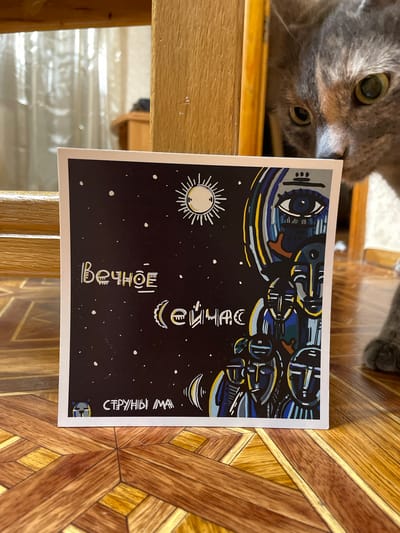 The image shows a close-up of a card with a stylized, cosmic-themed illustration on a dark background with white dots resembling stars. The illustration features a large eye with intricate patterns, a crescent moon-like shape with radiating lines, and abstract, possibly mechanical, forms in shades of blue, yellow, and grey. There is also a depiction of a sun with rays.

The card is partially obscured by a grey cat lying on its back, its head and front paws visible in the upper left portion of the frame. The cat's eyes are open, looking towards the card.

The card is placed on a wooden surface with a patterned inlay of light and dark brown wood. The lighting suggests it is daytime. Visible text on the card appears to be in Cyrillic script. The text reads "ВЕЧНОЕ" (Vechnoye, meaning "eternal") vertically on the left side of the card and "СТРУНЫ ВОЛНА" (Struny Volna, meaning "strings of the wave") vertically on the right side, in a stylized font that complements the illustration.
