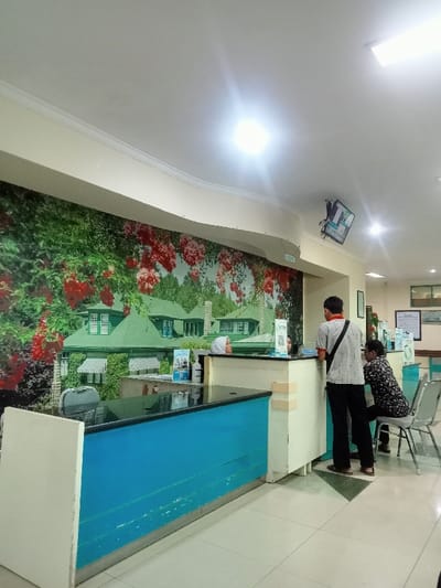 The photo shows a waiting area in a medical facility. A reception desk with a light blue panel sits in the foreground, manned by a person wearing a hijab. A man in a gray shirt is standing and talking to her, while a woman is seated on a metal chair nearby. The wall behind the desk has a large mural depicting a house surrounded by flowers. A television screen is mounted on the ceiling. The floor is light-colored tile.