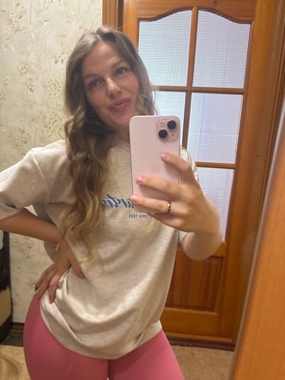 A young Caucasian woman with long, wavy blonde hair and blue eyes is taking a mirror selfie in a room. She is wearing a light gray sweatshirt with blue lettering and bright pink leggings. She has her right hand on her hip and her left hand holding a pink smartphone, taking the picture. A ring with small stones is visible on her left ring finger. The background includes a floral patterned wallpaper, a wooden door with frosted glass panels, and a wooden door frame. The overall setting appears to be a home interior. There is no information available about the location in the city of Kropyvnytskyi, Ukraine.