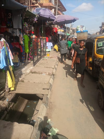 The image captures a bustling street scene in Fegge, Nigeria, likely a market area. On the left, a clothing stall displays a variety of garments, including t-shirts with graphics and colorful fabrics, hung from racks and shelves. Various umbrellas, some damaged, provide shade over the stalls. The ground is paved with concrete slabs, and a partially covered drainage ditch runs alongside the sidewalk.
A group of people are walking on the street. In the foreground, a woman in a black dress walks towards the viewer, carrying a brown crossbody bag. Behind her, a man in a black t-shirt and another person in a black and white checkered shirt are walking. Further back, several other individuals can be seen, some carrying items. A yellow auto-rickshaw, common in Nigerian cities, is parked on the right side of the street. The overall environment suggests a typical day in a busy Nigerian marketplace, with vendors, shoppers, and a rickshaw contributing to the vibrant atmosphere.