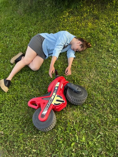 A young woman with her hair in a bun is lying on her back in the grass, appearing to be asleep. She is wearing a light blue button-up shirt and a dark gray skirt, paired with black platform shoes. Her right arm is extended towards a red toy motorcycle with black wheels, which is lying on its side next to her. The toy motorcycle has yellow and black graphics. The scene is set on a grassy lawn, likely outdoors, with dappled sunlight suggesting it is daytime. The overall mood appears peaceful and relaxed. The location context is identified as Мещёры, Russia. There is some visible text on the toy motorcycle, appearing to be "MX ON".