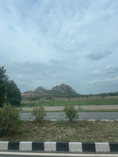 A scenic view captures a road passing through a landscape with rocky hills and green fields under a cloudy sky. In the foreground, the edge of a road is visible with a black and white concrete divider. Beyond the divider, grass and some small bushes grow. A two-lane road stretches horizontally across the frame, with a motorcycle parked on the side. In the middle ground, green fields are cultivated, and trees dot the landscape. The background is dominated by a range of rugged, rocky hills. The sky is overcast with scattered clouds. The overall scene appears to be from the outskirts of Addakal, India, a region known for its natural beauty and agrarian settings.