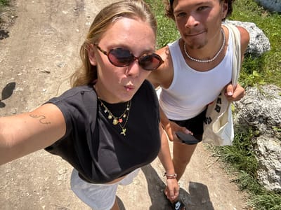 Two people are in the frame, likely a couple. One individual is wearing a white tank top and a pearl necklace. The other person has on a black t-shirt and sunglasses and is taking a selfie. They are standing on a paved ground with vegetation to the left and gravelly stone-like ground to the right. Both appear to be holding hands. A tote bag is slung over the shoulder of the individual in the white tank top. The location appears to be outdoors and in Sainte-Anne, France.