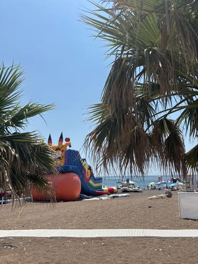 This image depicts a beach scene in Boreti, Montenegro. A large, colorful inflatable slide, designed with a character resembling Spongebob Squarepants, is set up on the dark sand. Inflatable rafts and other beach equipment are scattered around. The sea is visible in the background, with a few boats on the water. Several beachgoers are present, some under umbrellas. Palm trees frame the scene, with their fronds casting shadows. The sky is clear and blue. A white structure with some signage is visible on the right side of the image.
