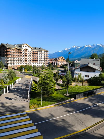 The image depicts a view of a resort area with a large, multi-story building, likely a hotel or apartment complex, on the left side of the frame. This building features a facade with numerous balconies and a predominantly beige and brown color scheme, set against a clear blue sky. In the middle ground, there are several smaller buildings, including one with a white facade and a dark, sloped roof. The area is characterized by lush green grass, scattered trees, and a paved road with a pedestrian crossing marked with yellow and white stripes. A lamppost stands near the road. In the background, a range of snow-capped mountains under a bright blue sky provides a scenic backdrop. The scene is well-lit, suggesting it was taken during daylight hours. The overall impression is that of a well-maintained urban or semi-urban resort environment in a mountainous region, consistent with locations like Estosadok, Russia. There are no people visible in the image.