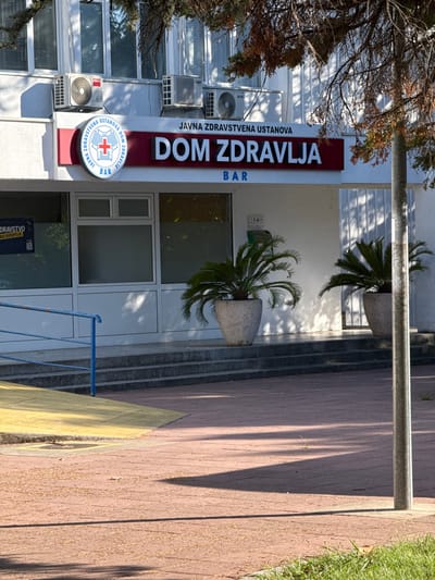 The image shows the exterior of a public health institution named "DOM ZDRAVLJA" in the city of Bar, Montenegro. The building is white with several windows and air conditioning units visible on the facade. A red sign above the entrance displays the name of the institution in large white letters, with smaller text above it and a circular logo to the left. The logo is blue and white with a red cross. Below the main sign, the word "BAR" is visible. In front of the building, there are two potted plants and a set of steps leading to the entrance. A yellow ramp is also visible on the left side, and a metal pole stands in the foreground on the right. The ground in front of the building is paved with reddish-brown tiles, and some dry leaves are scattered on the ground. The overall scene appears to be a daytime shot with sunlight casting shadows.