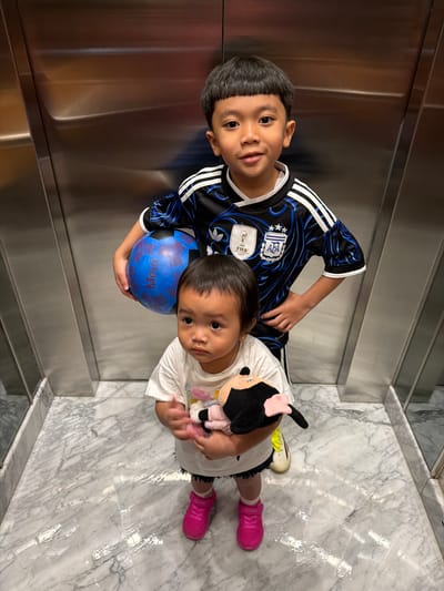 Two children, a boy and a younger girl, stand in an elevator with brushed metal walls and a marble floor. The boy, wearing a black and blue soccer jersey, holds a blue soccer ball with red markings. The girl, in a white t-shirt, clutches a stuffed toy. The scene appears to be an indoor location in Central Jakarta, Indonesia, likely within a residential or commercial building.