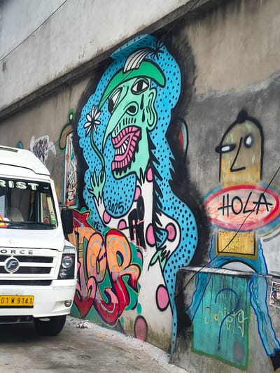The image shows a street art mural on a concrete underpass. The mural features a cartoonish face with its mouth open and pink tongue, stylized dragon-like figures in blue and green, and abstract colorful shapes. There is text that reads "HOLA" within a red and yellow oval on the upper part of the mural. A year, "2025", is visible within the blue dragon-like figure. The overall style is vibrant and whimsical.

Below the mural, the front of a white vehicle, likely a truck or van, is visible. The vehicle has a license plate that reads "01 W 9141". Text on the side of the vehicle appears to read "FORCE". The setting appears to be a public space, possibly a road or parking area under a bridge, given the concrete structure and the presence of the vehicle.

The lighting suggests it is daytime, and the weather is not discernible from the image. The graffiti art implies an urban or semi-urban environment. The art itself conveys a sense of playful energy and creativity. The overall impression is of a public space decorated with dynamic and colorful graffiti art, with a vehicle parked in front of it.
