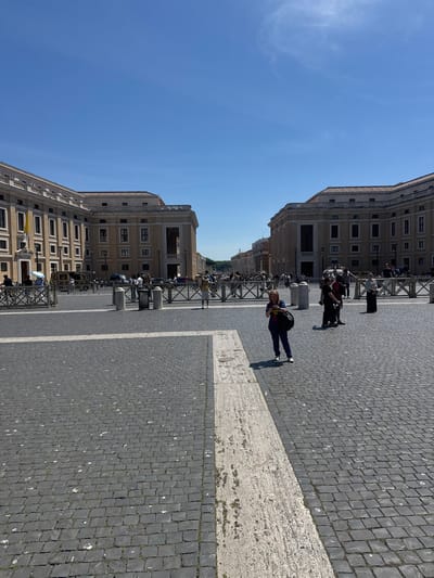 The image captures a bright day in St. Peter's Square, Vatican City, Rome, Italy. The sky is a clear blue. The square is paved with dark cobblestones, marked by a wide, light-colored border. Buildings with classical architecture line the square. Numerous people are present, some standing behind metal barriers. A woman in blue jeans and a dark shirt stands in the foreground.