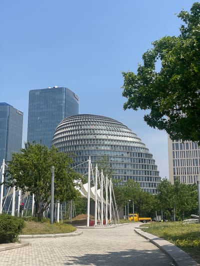 The image captures an urban scene in the Chaoyang District of China, featuring a mix of modern architecture and green spaces. A prominent, dome-shaped building with a grid-like facade stands among taller, rectangular skyscrapers. Trees partially obscure the view, adding natural elements to the cityscape. A paved walkway runs along the right side of the frame, suggesting a pedestrian area. In the background, there is a yellow truck. A traffic light is also visible. White structures, possibly part of a public art installation or architectural feature, are seen near the walkway. The sky is clear and blue, indicating a sunny day.