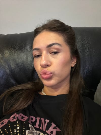 A young woman with long, dark brown hair, wearing a black t-shirt with pink lettering and sequins, is shown in a close-up selfie. She is leaning back against a black leather couch, with a light gray wall in the background. The woman is making a kissing face, with her lips pursed. She has light makeup on, including blush and mascara. The image appears to be taken indoors. The location of Novi Iskar, Bulgaria is not discernible from the image itself.