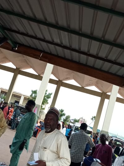 The image depicts an outdoor gathering, possibly a market or a transportation hub, in Gombe, Nigeria. The foreground features several men, predominantly wearing traditional attire. One man in the center is wearing a cream-colored kaftan and a cap, holding a stack of papers. To his left, another man is dressed in a green kaftan. In the background, a crowd of people are visible, along with several vehicles, including vans and cars. The architecture suggests a covered waiting area or market stall, with a corrugated metal roof supported by concrete pillars. The overall atmosphere appears busy and communal.