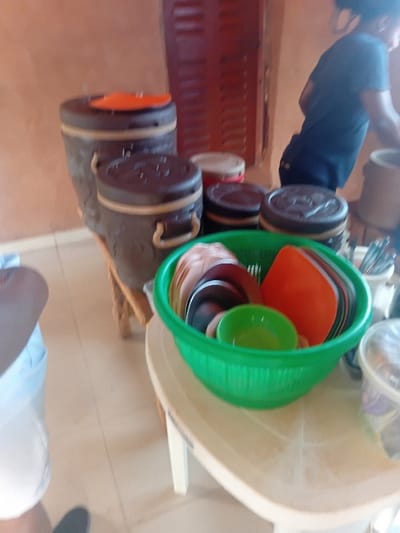 This image depicts an indoor scene within a commercial establishment, likely an eatery or shop, in Ejule, Nigeria. The foreground shows a light-colored tiled floor. On the left, the lower torso and legs of an individual wearing a light-colored top and dark footwear are partially visible. A light-colored, possibly beige or off-white, plastic table occupies the mid-ground, holding a green plastic colander filled with various dishes, including cream-colored plates, a dark plate, a small green bowl, and an orange rectangular item. To the right of the colander, a transparent plastic container with a lid is visible. Behind the table, several dark brown cylindrical insulated containers or food warmers with lighter bands and handles are arranged; one taller unit has an orange item on its lid. In the background, a person wearing a dark blue t-shirt is partially visible from the waist up, their head and upper body obscured as they appear to be leaning or reaching towards an object out of frame to the right. The background wall is a light brown or orange hue, featuring a prominent reddish-brown wooden louvered shutter or window.