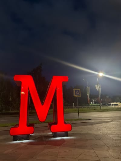 A large, red, illuminated letter "M" stands prominently on a paved area at night. The letter is a three-dimensional structure, likely a sign, with internal lighting that casts a glow. In the background, there is a road, a pedestrian crossing sign, lampposts with flags, trees, and a building with windows. A white bus or van is partially visible on the road in the distance. The sky is dark, indicating it is nighttime, with some clouds and light streaks from artificial lighting. The ground is paved with geometric stone tiles. The scene appears to be an outdoor public space, possibly a street or a plaza, in Naberezhnye Chelny, Russia.