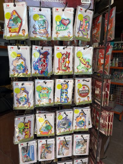 The image shows a retail display of souvenirs in Nusa Penida, Indonesia. The souvenirs are primarily magnets, most of which feature the "I Love Indonesia" logo. The magnets are packaged in clear plastic and hung on black metal display racks. Designs on the magnets include scenes of beaches, the text "Diamond Beach," "Nusa Penida," and "Bali." Some magnets have Japanese text that translates to "Glow in the dark." Several of the magnets are shaped like dinosaurs or cartoon characters. The display also includes some flat souvenir cards with similar designs. In the background, shelves stocked with other products, likely more souvenirs or general store items, are visible, along with a partial view of an orange shopping cart. The lighting suggests an indoor setting, possibly a gift shop.