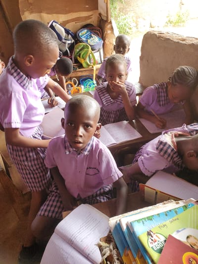 The image captures a group of young children engaged in a learning activity within what appears to be a simple classroom in Ukwu-Abwa, Nigeria. Several children are visible, mostly wearing light purple shirts with plaid collars and cuffs, indicative of school uniforms. Their hair is closely cropped or styled in braids.
The children are seated at desks or benches, diligently working in notebooks with pencils. One child in the foreground looks directly at the camera with an open expression, while another nearby covers their mouth. Stacked books, some with colorful covers, are visible on a desk to the right, with one clearly showing "Friday School" and "EDGE" on its cover. School bags and what appear to be colorful learning toys or aids are scattered in the background, suggesting a dynamic learning environment.
The setting features rough, unadorned walls, possibly made of stone or concrete, and natural light filters in from an unseen opening, indicating it is daytime. Outside, some greenery is visible, adding to the rustic feel of the location. The overall atmosphere suggests focused learning despite the simple surroundings.
Visible text includes "WATESSOR SCHOOL" and "NOWELLE EZLINNAKA" on a child's uniform. In open notebooks, various handwritten lines are discernible, including phrases like "Risks are Ring...", "COWISE SOMEtting bad to happen...", "Something a risk factor," "pre existing," and "Look and serve Foo." There are also numbers like "260126" and words like "sefe" and "Friday Sochina" (likely "School").