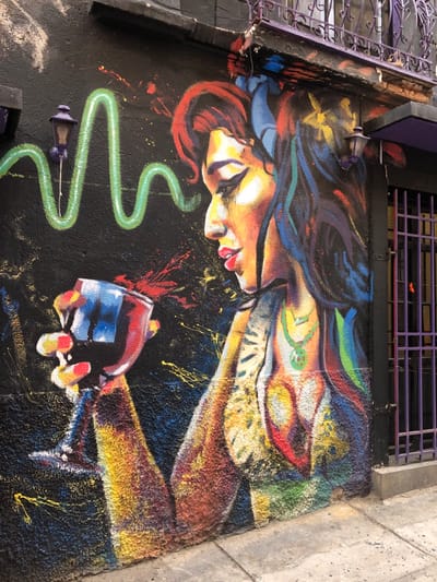 A vibrant, colorful mural features a woman lying horizontally, her face illuminated with warm tones, framed by swirling hair in shades of red, blue, and black. She seems to be holding a glass, painted in bright red, blue, and white, above a textured black background. A lime-green, serpentine shape meanders near the glass, punctuated by small, purple fixtures. The painting is positioned on a rough surface, flanked by a light-grey wall and a dark-grey iron structure.