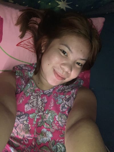 A young woman with brown hair and fair skin is lying down on a pink pillow with a floral pattern. She is wearing a pink top with a floral pattern as well. She is smiling at the camera and her eyes are open. The background is dark, possibly a bedroom. The image appears to be a selfie taken by the woman. The image is not located in the city of Kiblawan, Philippines.