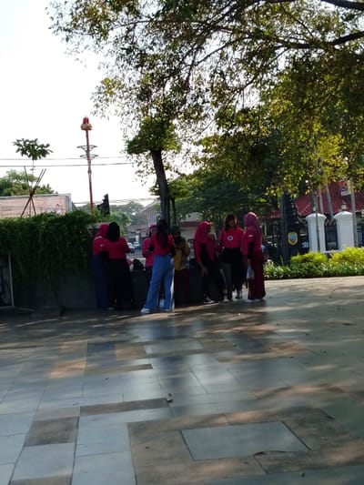 A group of nine people, all wearing pink or red shirts, stand in a paved area in front of a building. The people are mostly women, with one male individual. Most of the women are wearing headscarves. The group is standing in a semi-circle formation. There are a few trees in the background, with a tall red structure in the distance. The pavement is a light gray color with a pattern of squares. The area is located in Pati, Indonesia, at the coordinates latitude: -6.753, longitude: 111.04.