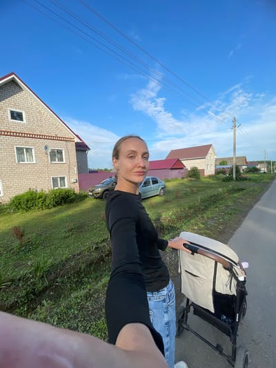 A woman, facing the camera with her right side angled away, walks down a paved road in a rural setting in Podshivalovo, Russia. She is pushing a beige and black stroller with her right hand. She wears a black long-sleeved shirt and blue jeans. Behind her, to the left, is a two-story brick house with a red roof. A green car is parked in front of a purple fence to the right of the house. Further down the road, more houses and a utility pole are visible under a blue sky with scattered clouds.